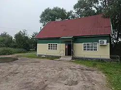 Здание с покатой крышей. Стены жёлто-зелёные, крыша кирпичного цвета. Перед крыльцом - площадка без дорожного покрытия, рядом - большая лужа.