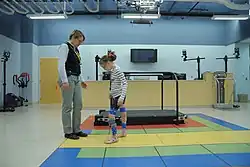 Девочка, носящая брекеты, идёт к женщине в тренажерном зале с беговой дорожкой, видимой на заднем плане.