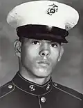 Head of a young Hispanic man wearing a dark military jacket and a white peaked cap with a black visor and an eagle-globe-and-anchor emblem on the front.