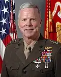 A color image of James Amos, a white male in his Marine Corps Service A uniform. He is not wearing a hat, several ribbons are visible as well as a rifle and pistol marksmanship badges and naval aviator insignia. The Marine Corps flag and United States flag are visible in the background.