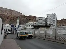 Эль-Мукалла. Маршрутные мини-автобусы на улице Старого Города. Слева от дороги, за забором и постройками море — в этом месте в середине XX века была старая гавань. За городом на вершинах гор видны три сторожевые башни.