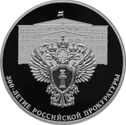 На переднем плане реверса монеты изображён герб, на фоне — здание Прокуратуры России в Москве