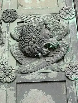 A relief with lion’s head facing right with bared teeth over four folded wings and a frame with floral bosses at each corner