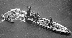 A large, damaged, stripped warship being hosed down by a smaller boat