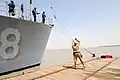 Британский матрос-швартовщик возвращает выброску на борт военного корабля ВМС США после закрепления продольного кормового швартова на пале пирса