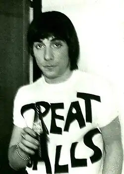 A cropped, black-and-white, and slightly out-of-focus photo of Keith Moon. Moon looks at the camera with a blank expression. He wears a white T-shirt with large lettering on the front. In his right hand he holds a bottle, the label of which is obscured by motion blur. He wears a ring on his right forefinger, and a metal bracelet on his right wrist. His hair is cut to shoulder-length, and has a fringe that reaches his eyebrows. Partially visible behind him is a white wall and an open door.