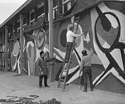 Karel Appel, 1955: Karel Appel is making a painting on the wall of an exposition-building in Rotterdam - photo, 19 April 1955 by Joop van Bilsen / Anefo - National Dutch Archive